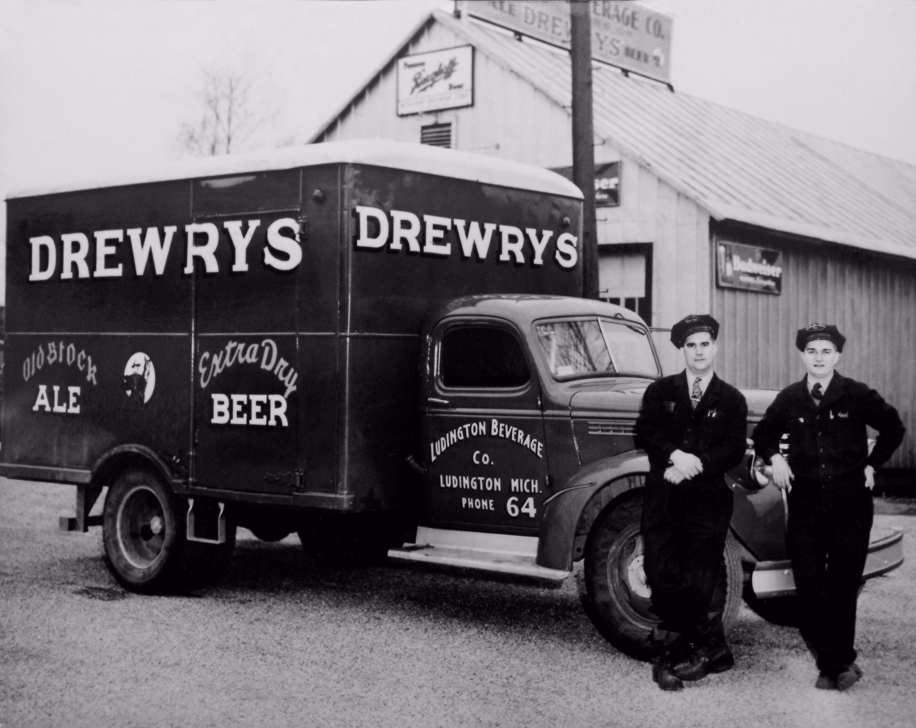 Two men in uniforms leaning against a vintage Drewrys beer delivery truck outside a warehouse.