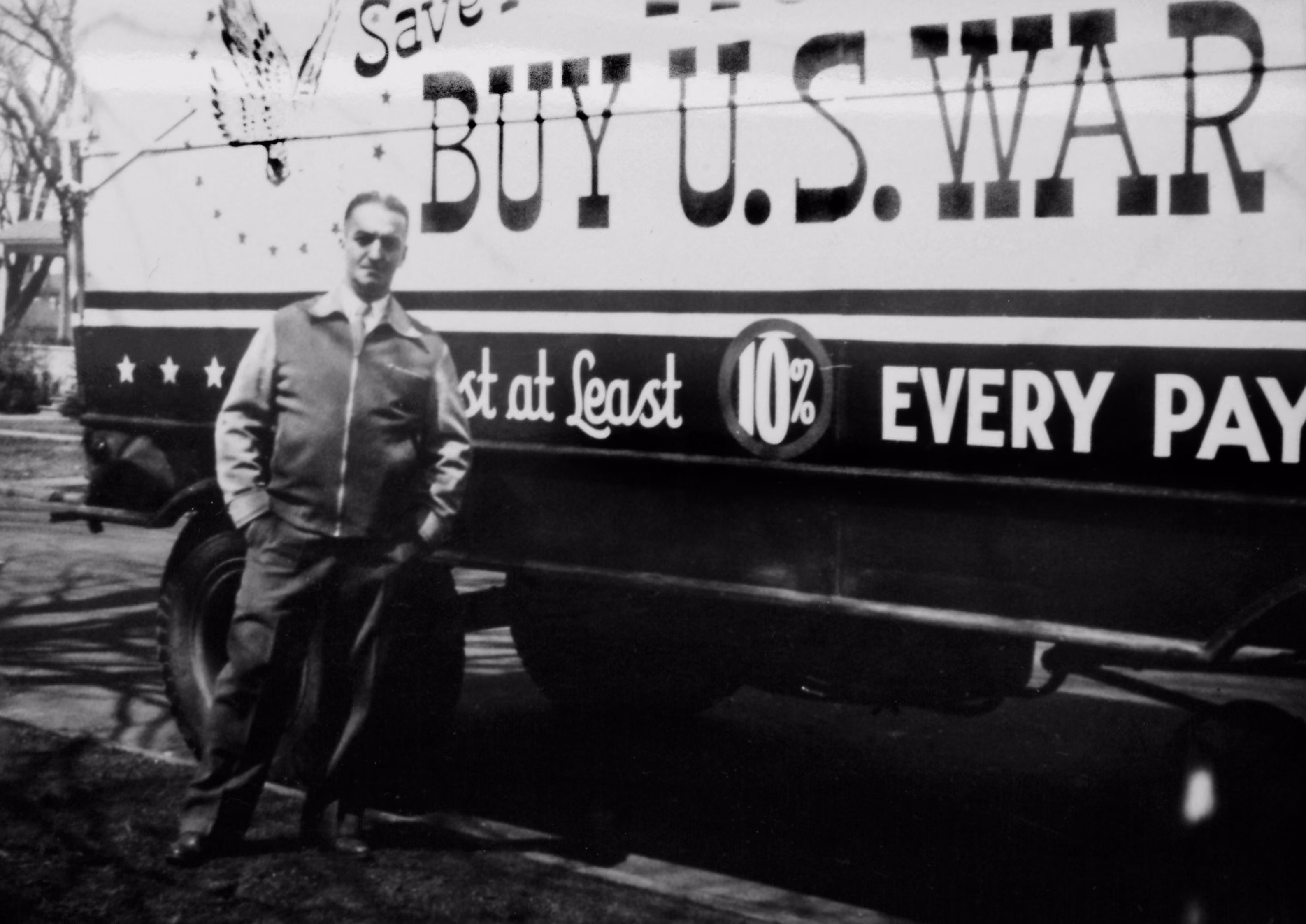 Man in jacket stands in front of a truck with a WWII-era 'Buy U.S. War Bonds' painted sign.
