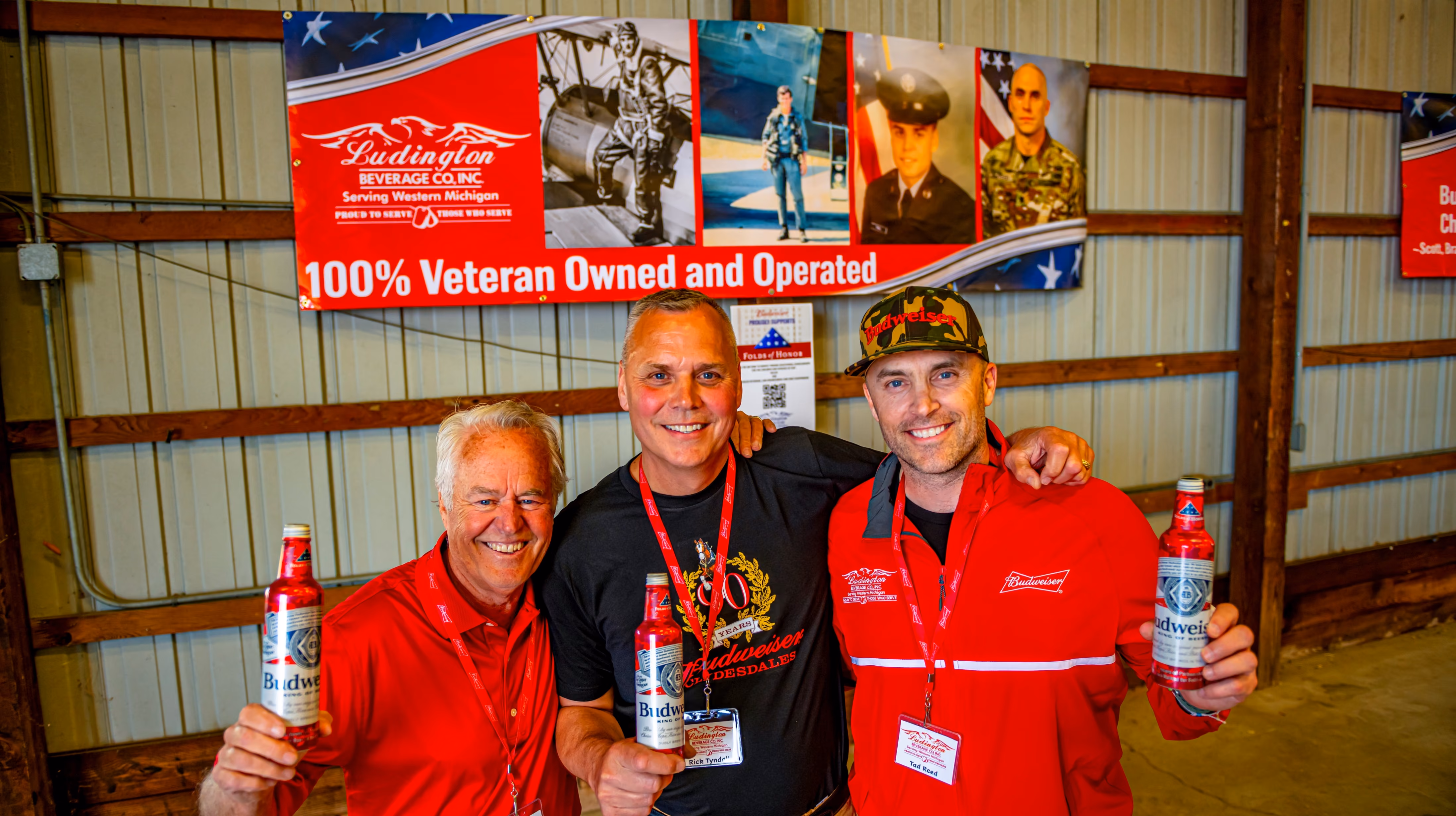 Three men smiling and holding Budweiser beers under a 100% veteran owned banner in a warehouse.