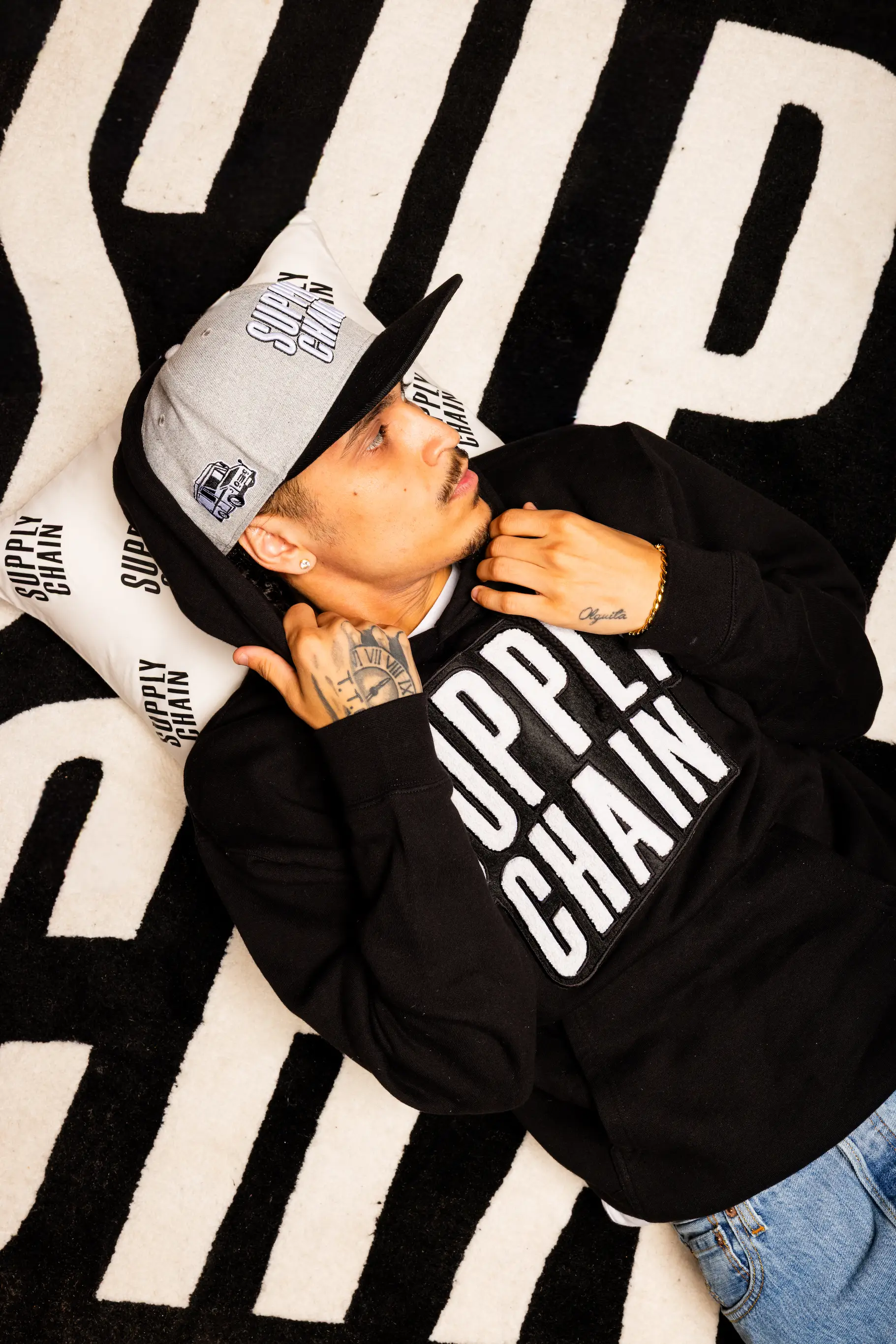 Model laying on Supply Chain Distro rug wearing logo hoodie — So & So Productions creative streetwear photo.