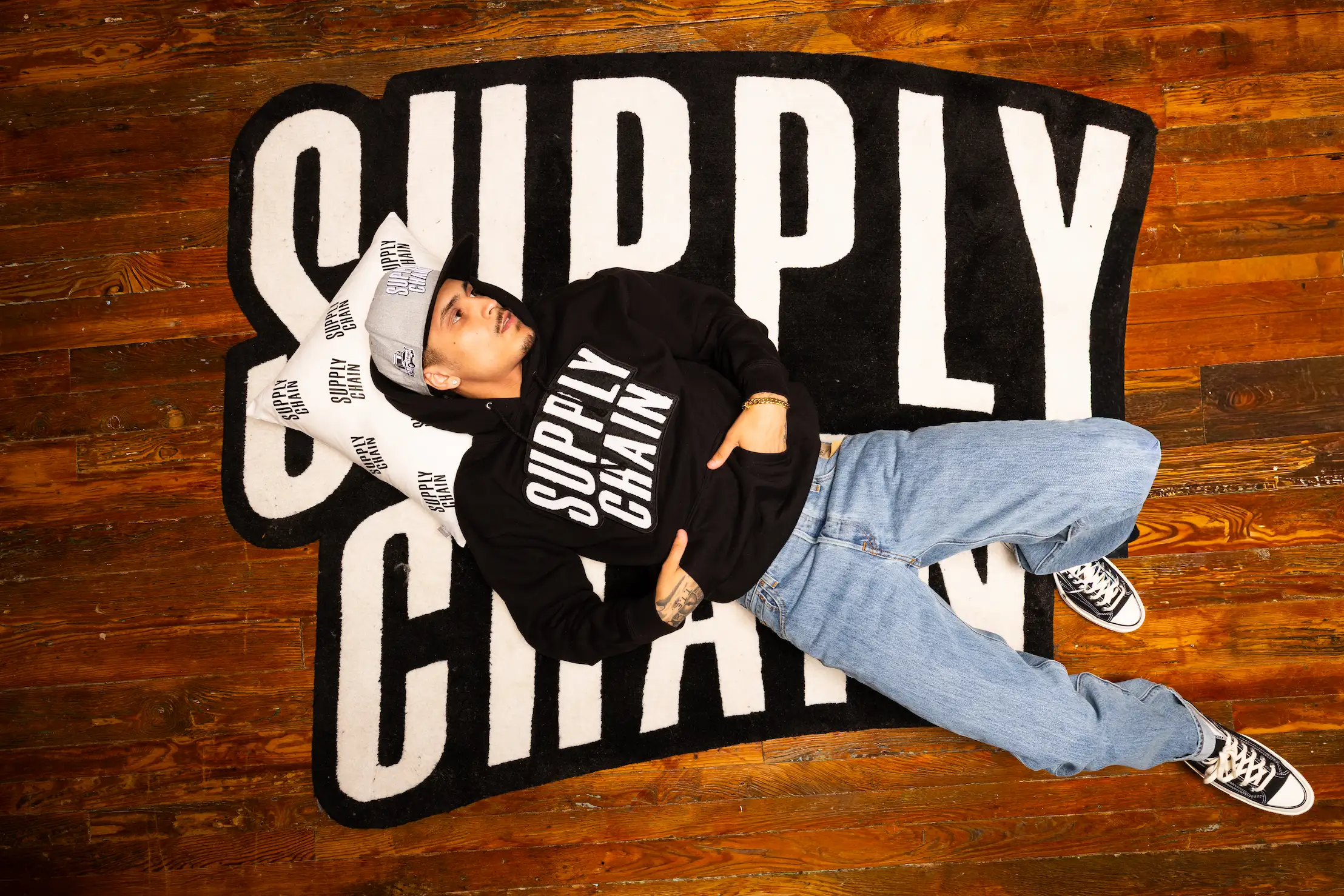 Candid overhead shot of model in Supply Chain Distro hoodie relaxing on branded rug — So & So Productions fashion campaign photography.