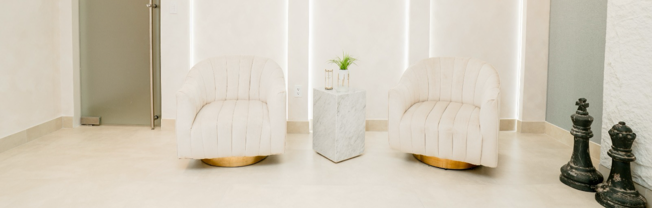 Minimalist waiting area with two beige upholstered chairs, a marble pedestal table with a plant and candle, and large chess piece statues.