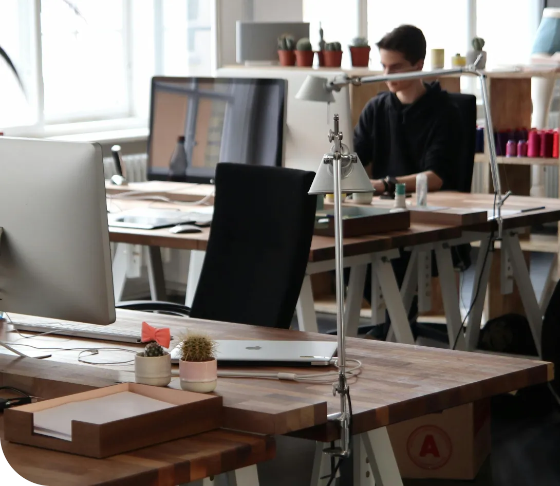 Modern office desk with computer, laptop, small potted cacti, and a lamp, with a person working in the background.