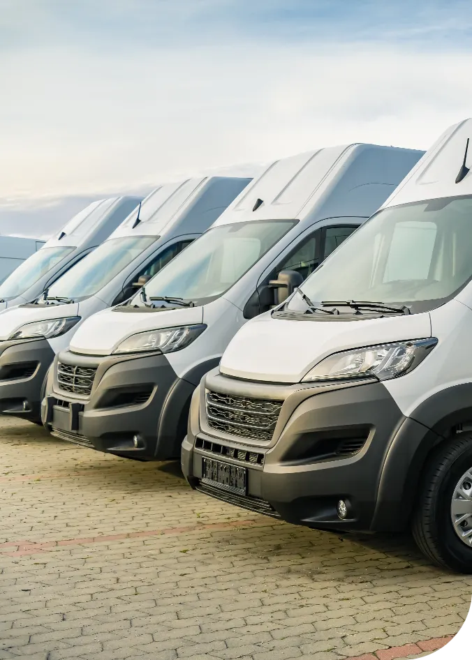 Row of white delivery vans parked side by side on a paved surface under a cloudy sky.