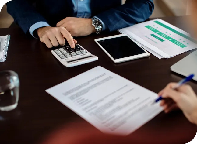 Person using a calculator while another person holds a pen over a document on a dark wooden table with a tablet, papers, and a glass of water.