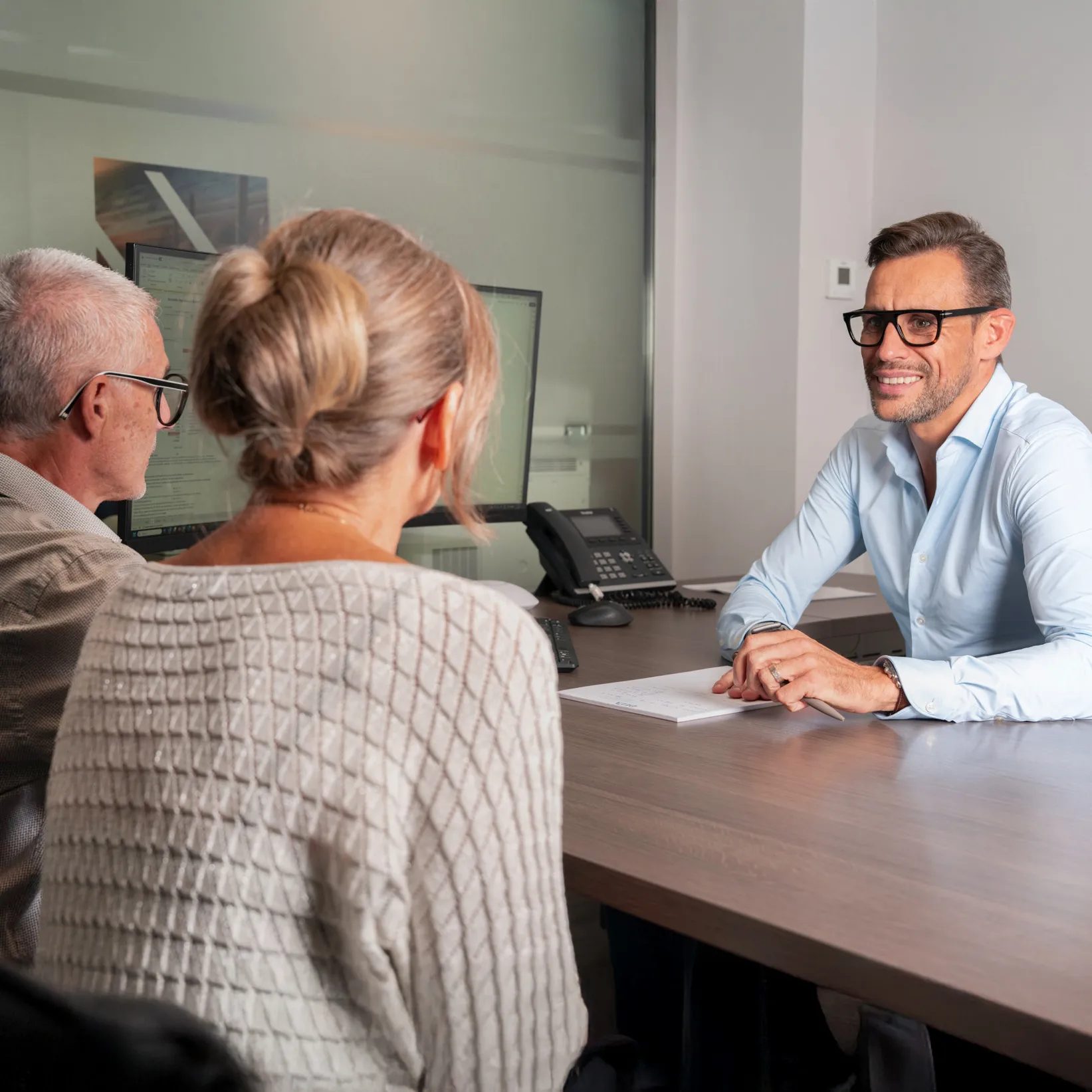 A man in glasses and a light blue shirt smiling and consulting with an older couple at an office desk.