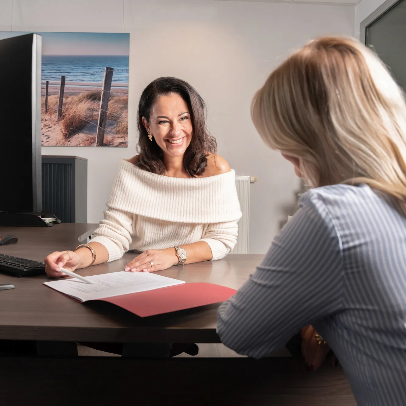Two women sitting across a desk in an office, one smiling and pointing to a document in a red folder.