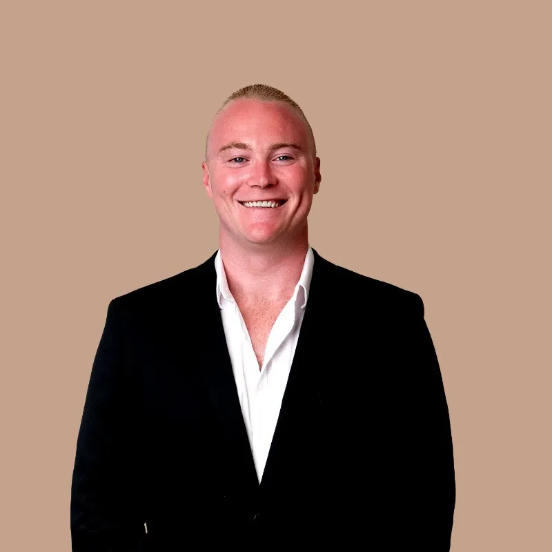 Smiling man with short blond hair wearing black suit jacket and white shirt against beige background.
