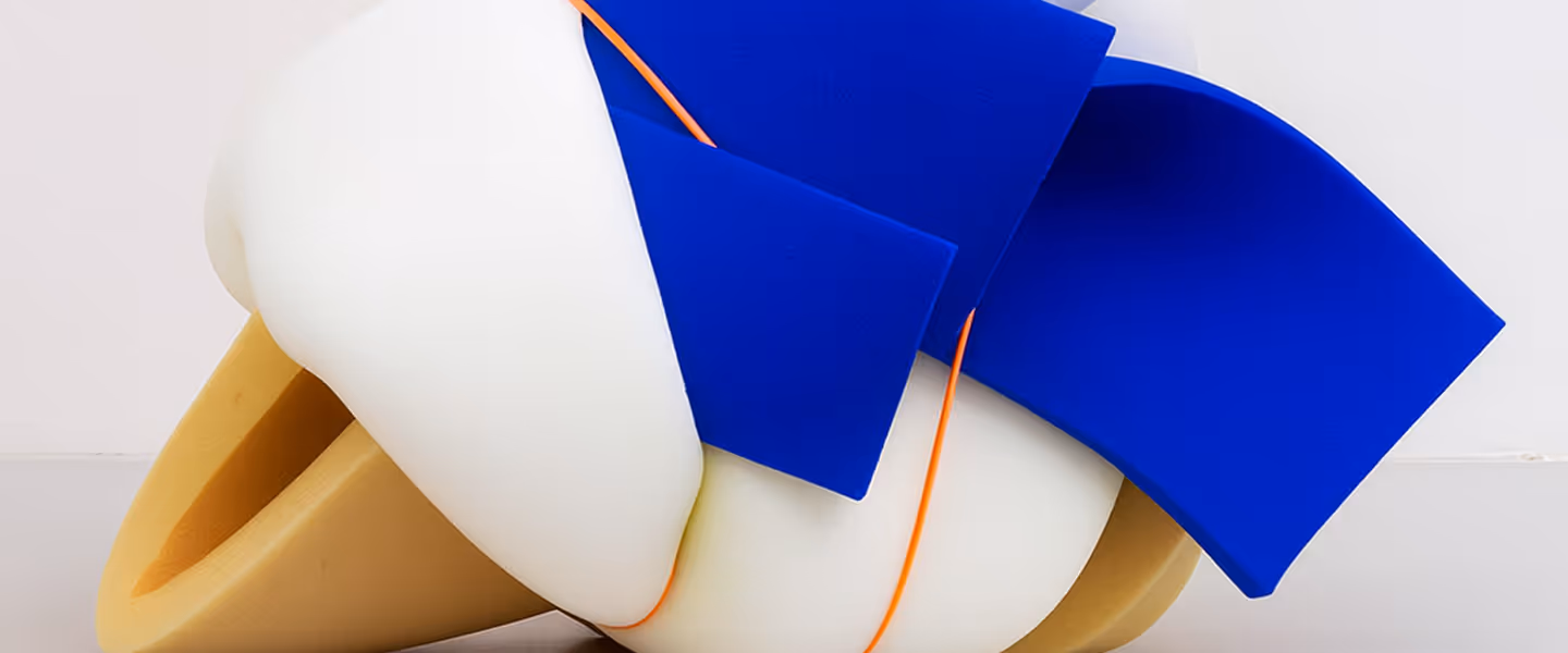 A soft, abstract sculptural object made of twisted white foam forms, wrapped with bright blue flat panels and thin orange bands, sitting on a neutral surface against a plain background.