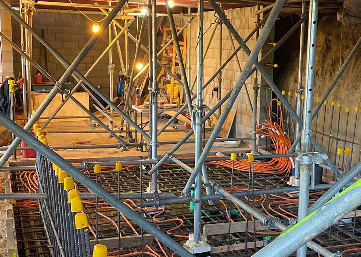 JLC construction site with metal scaffolding, rebar grid, and orange electrical conduits inside a concrete block structure.