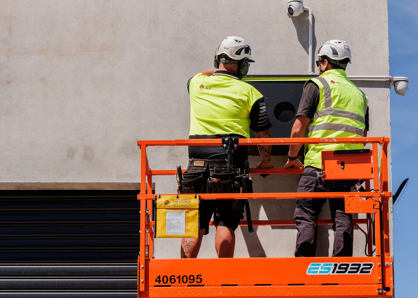 Two construction workers in safety helmets and yellow vests install equipment on a building wall from an orange elevated platform.