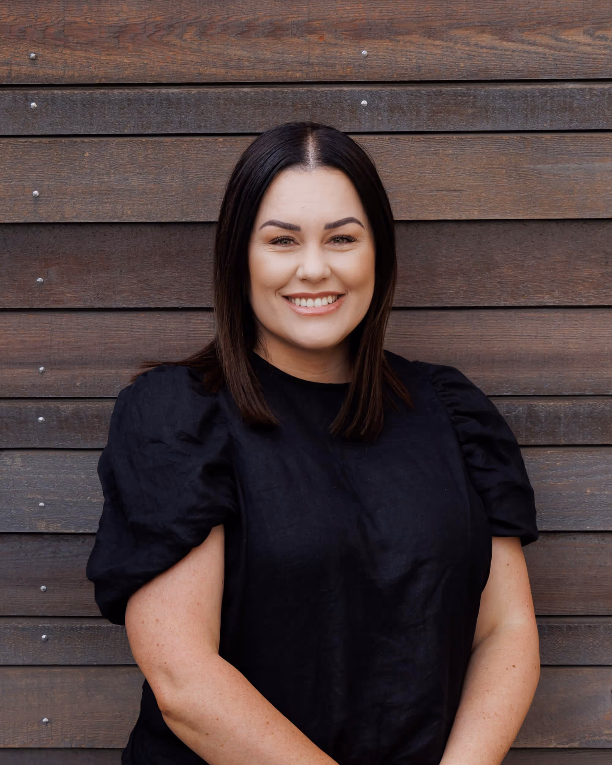 Tammy is the general manager at JLC she is a smiling woman with straight dark hair wearing a black puff-sleeve top standing against a wooden plank wall.