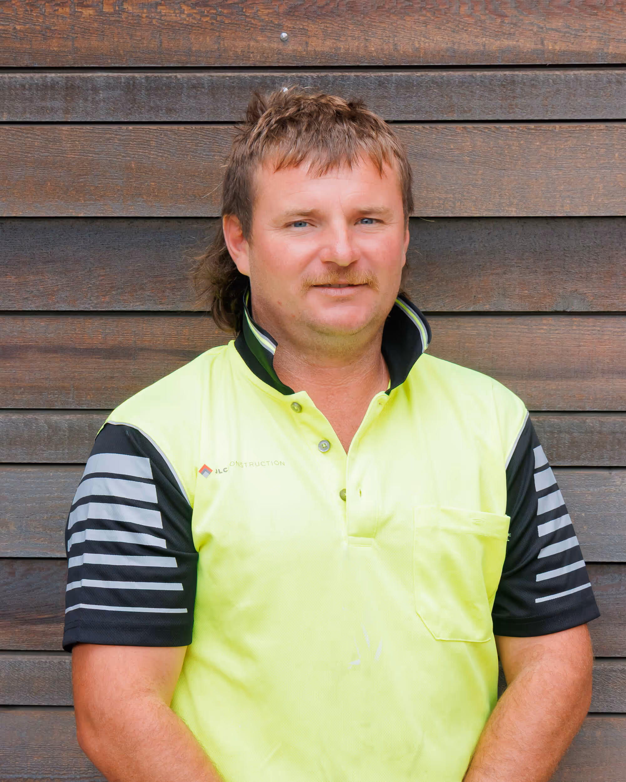 JLC construction worker called Aaron is a man with light brown hair and mustache wearing a neon yellow and black short-sleeve work shirt standing against a wooden wall.