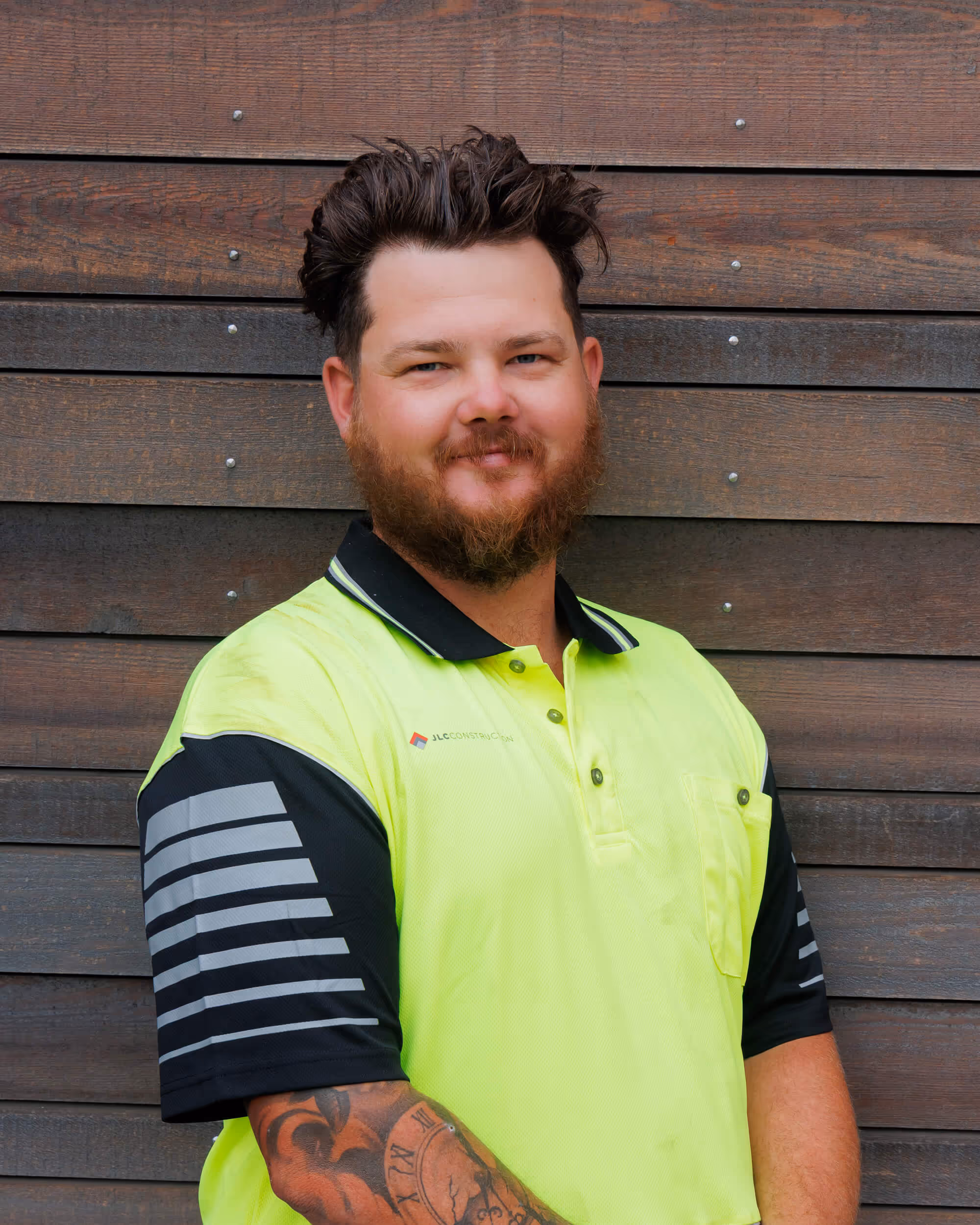 JLC construction worker called Alex is a man with beard and tattooed arm wearing a yellow and black polo shirt standing in front of a wooden wall.