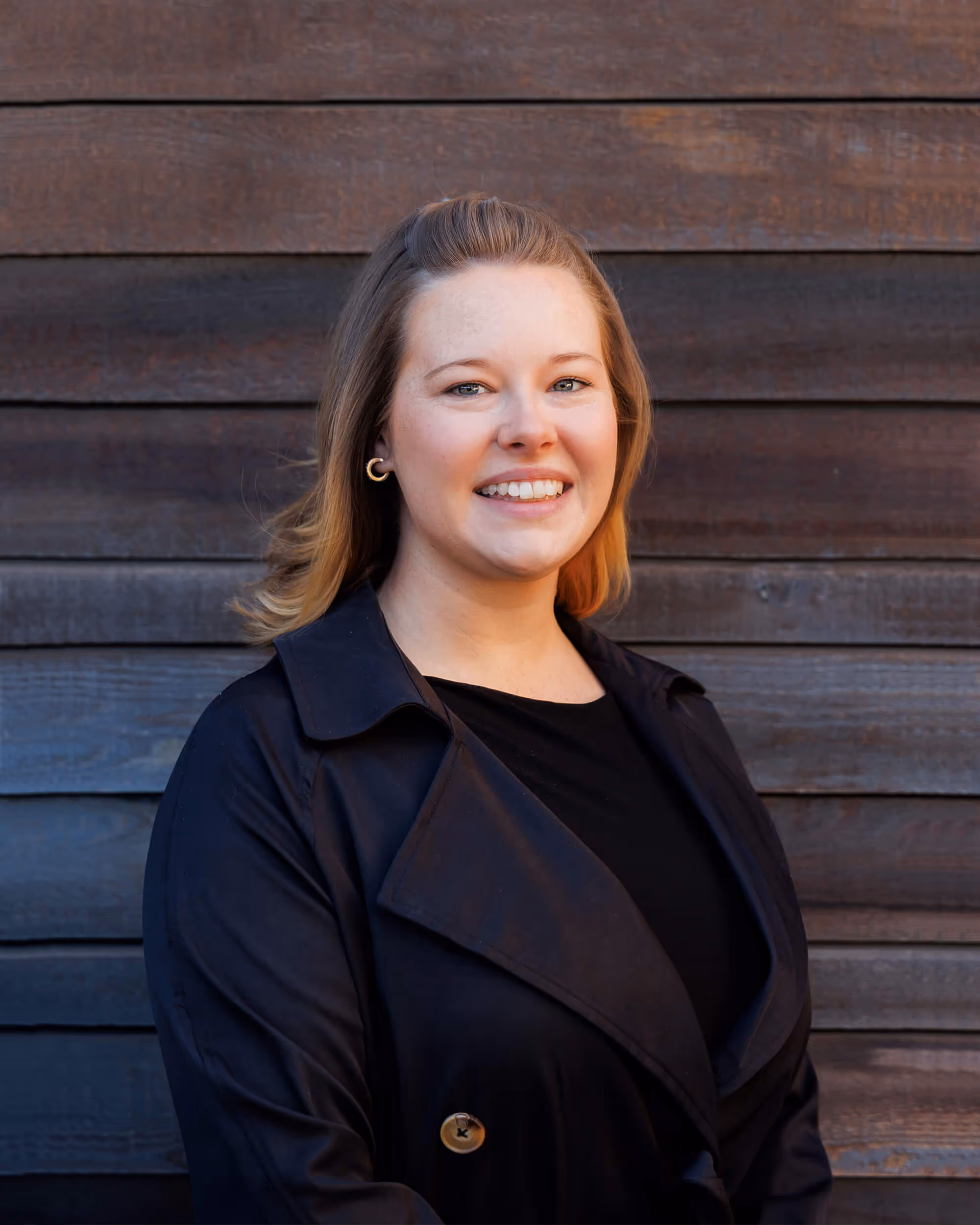 Jessica Milne, office manager at JLC is a smiling woman with blonde hair wearing a black coat standing in front of a wooden wall.