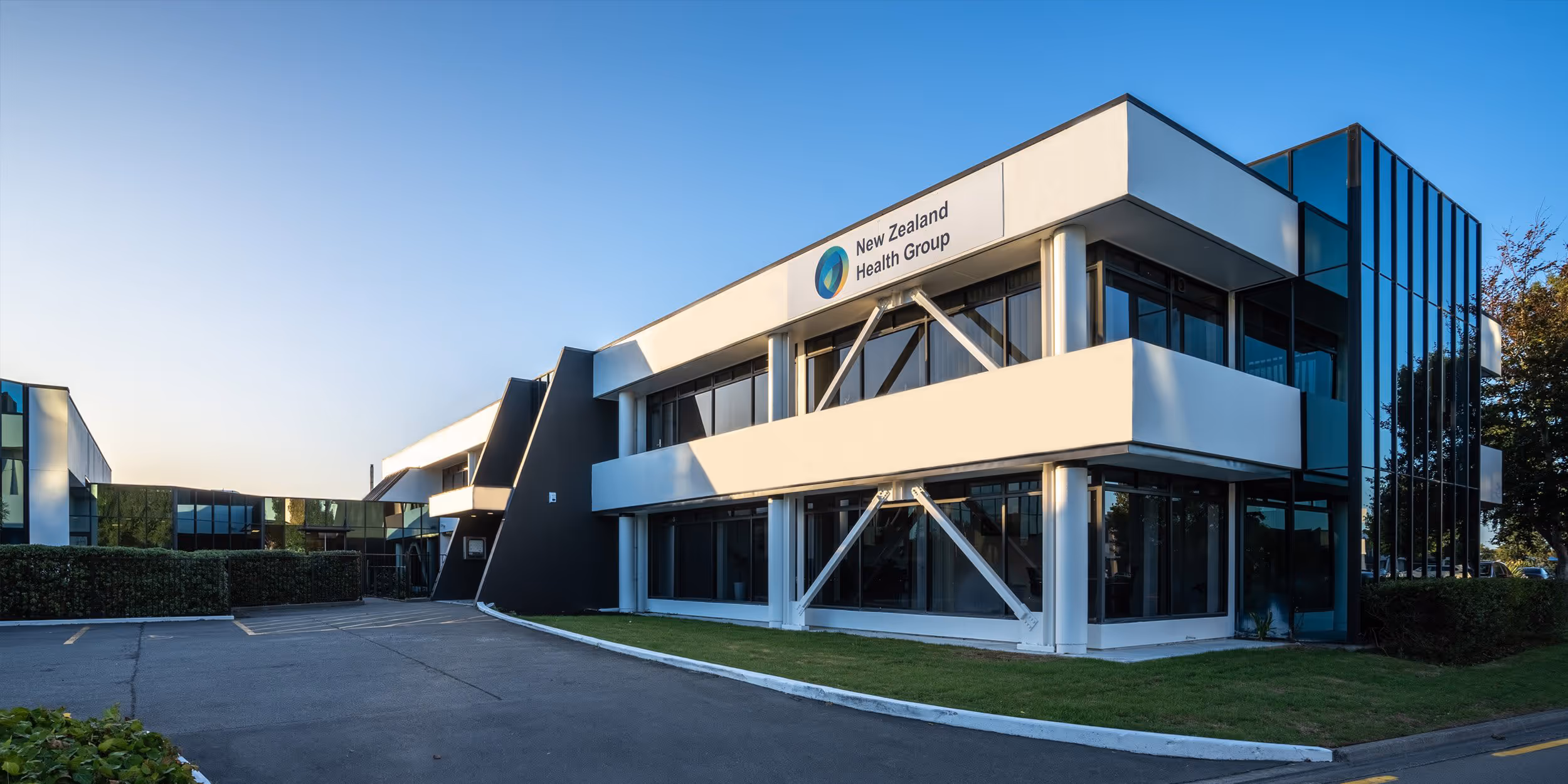 Modern two-story building with large windows and white exterior, labeled New Zealand Health Group, under clear blue sky.