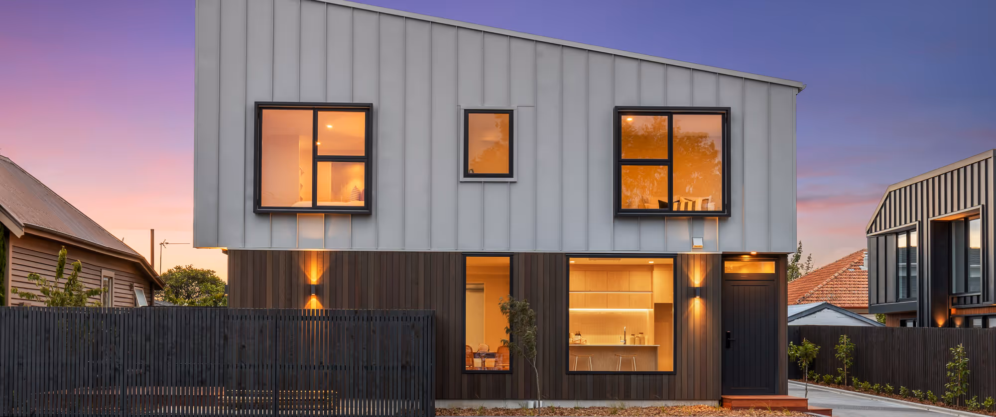 JCL Construction built this multi-unit modern two-story house with gray upper facade, dark wooden lower facade, large windows, and warm interior lighting at dusk.
