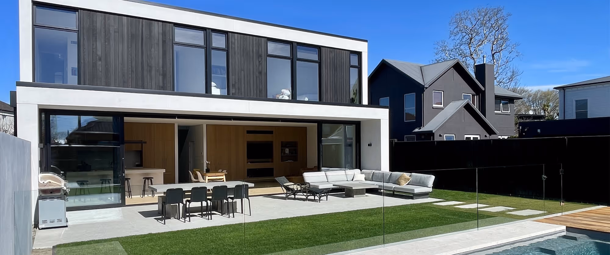 JLC Construction new build is a modern two-story house with large windows, open patio with dining table and lounge seating, glass fence, green lawn, and a pool.