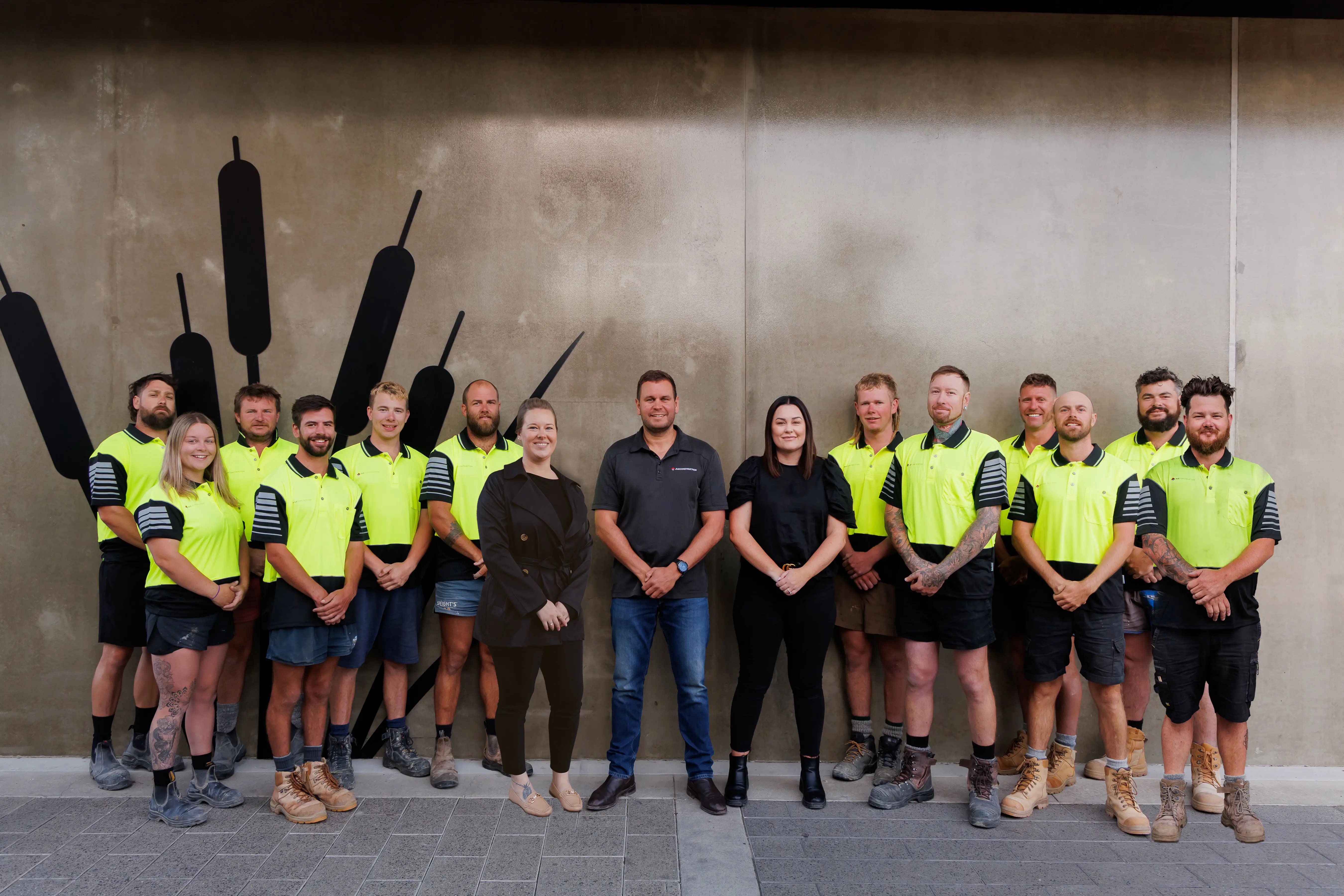 Group of JLC Construction staff standing in a row; most wear yellow high-visibility shirts with black shorts, and three wear casual black outfits, posing against a concrete wall with black abstract shapes.