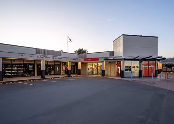 Exterior view of a commercial shopping centre in Fendalton, Christchurch, showing Jigsaw Hair salon and SuperValue supermarket with a large empty parking lot at dusk.
