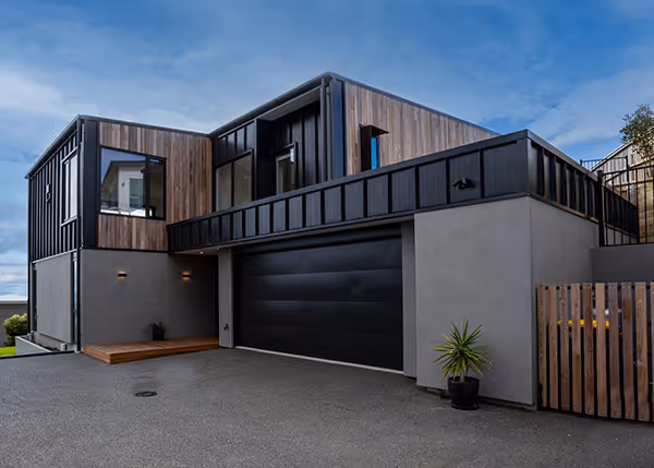 JLC Construction built this modern two-story house with black garage door, wood paneling, and large windows under a blue sky.