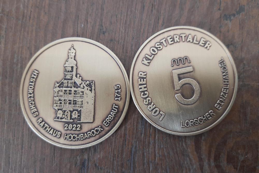 Zwei goldfarbene Medaillen auf Holzuntergrund, eine zeigt das historische Rathaus Hochbarock erbaut 1715, die andere den Schriftzug Lorscher Klostertaler mit der Zahl 5.