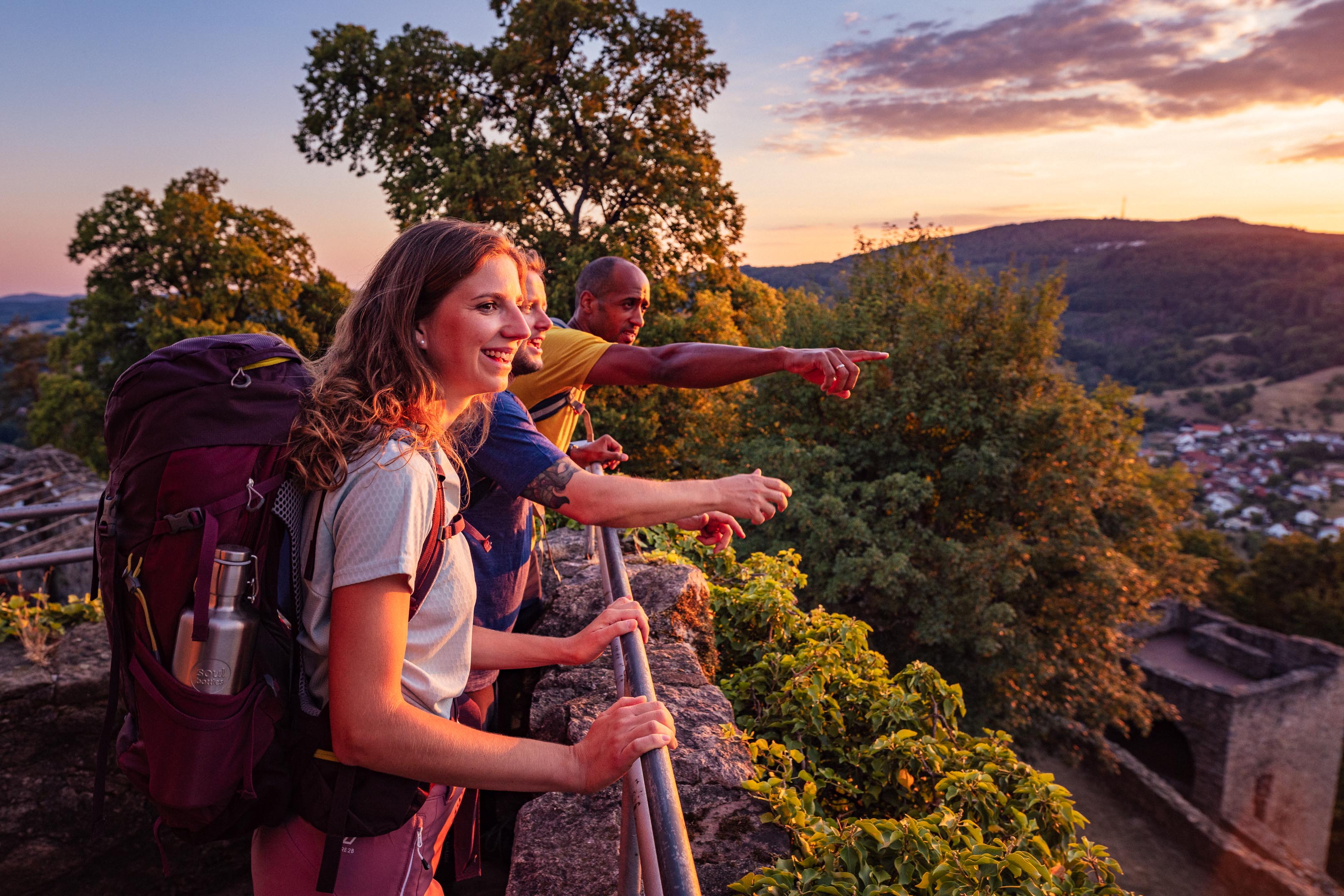 Drei Wanderer mit Rucksäcken genießen bei Sonnenuntergang die Aussicht von einer Aussichtsplattform auf bewaldete Hügel und ein Dorf.