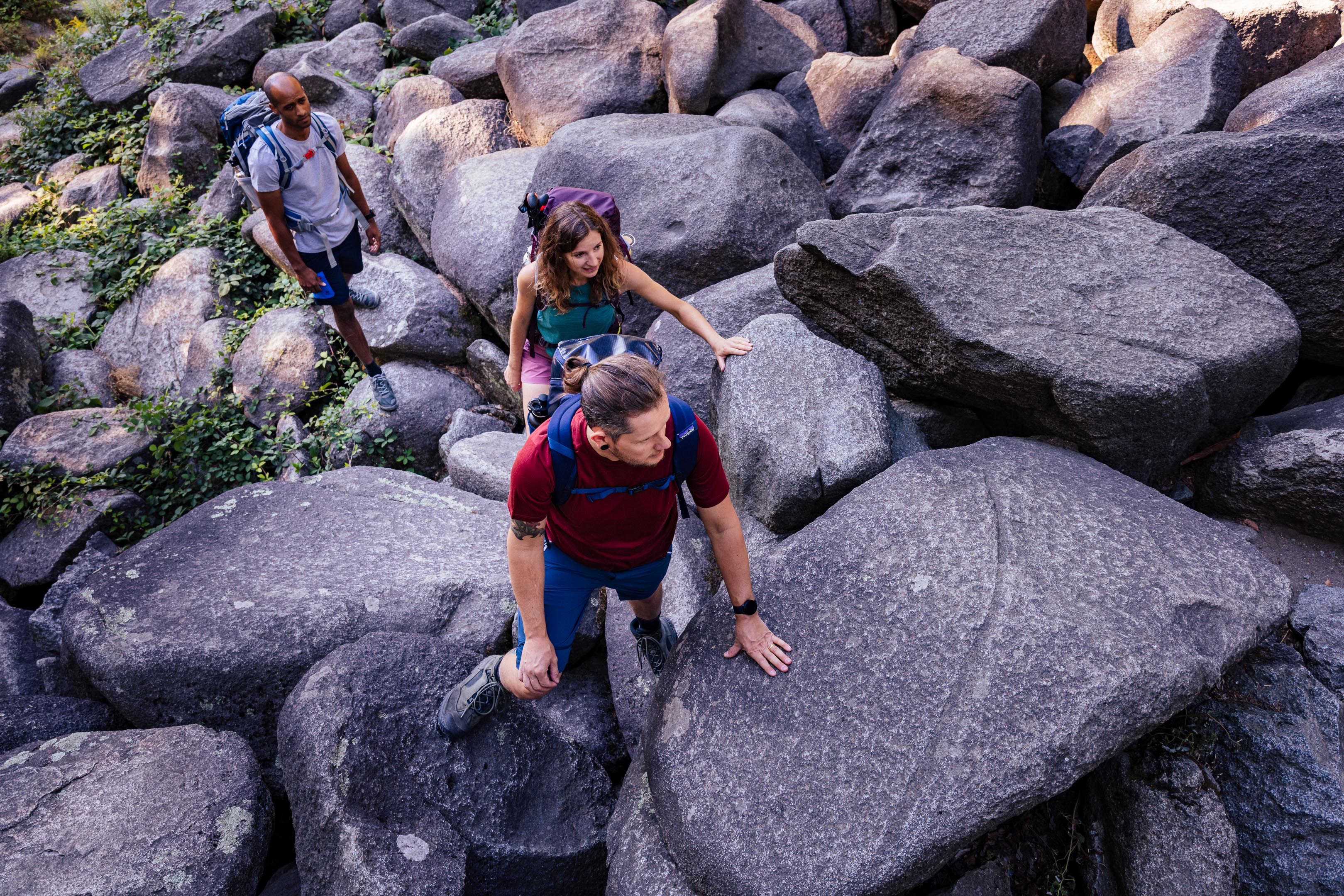 Drei Wanderer klettern über große graue Felsen auf einem felsigen Wanderweg.