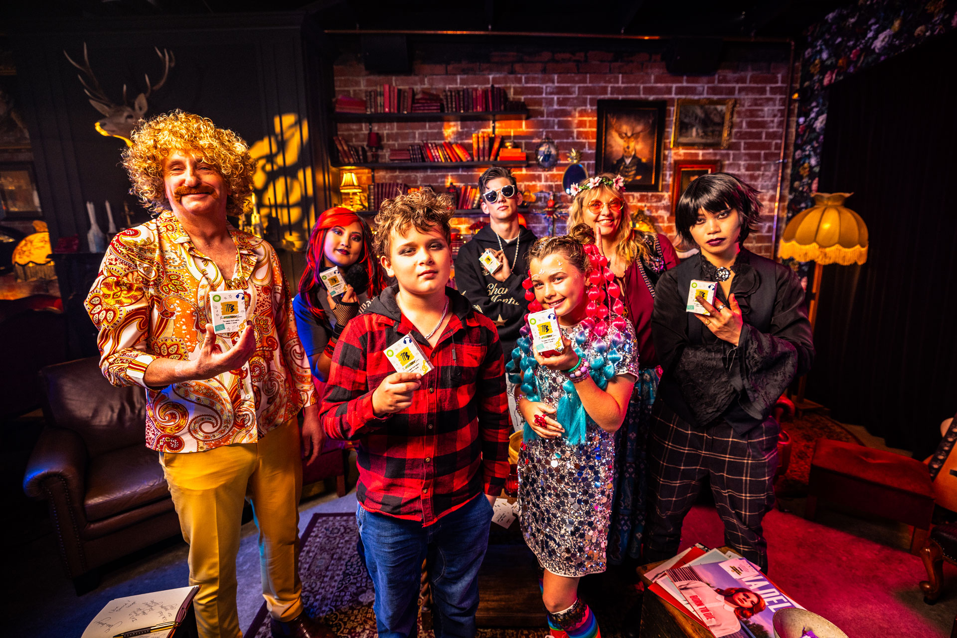 Group of seven people dressed in colourful and eclectic music genre-inspired clothes posing indoors, each holding a deck of The Playlist Game, with a cozy room featuring bookshelves and retro decor in the background.