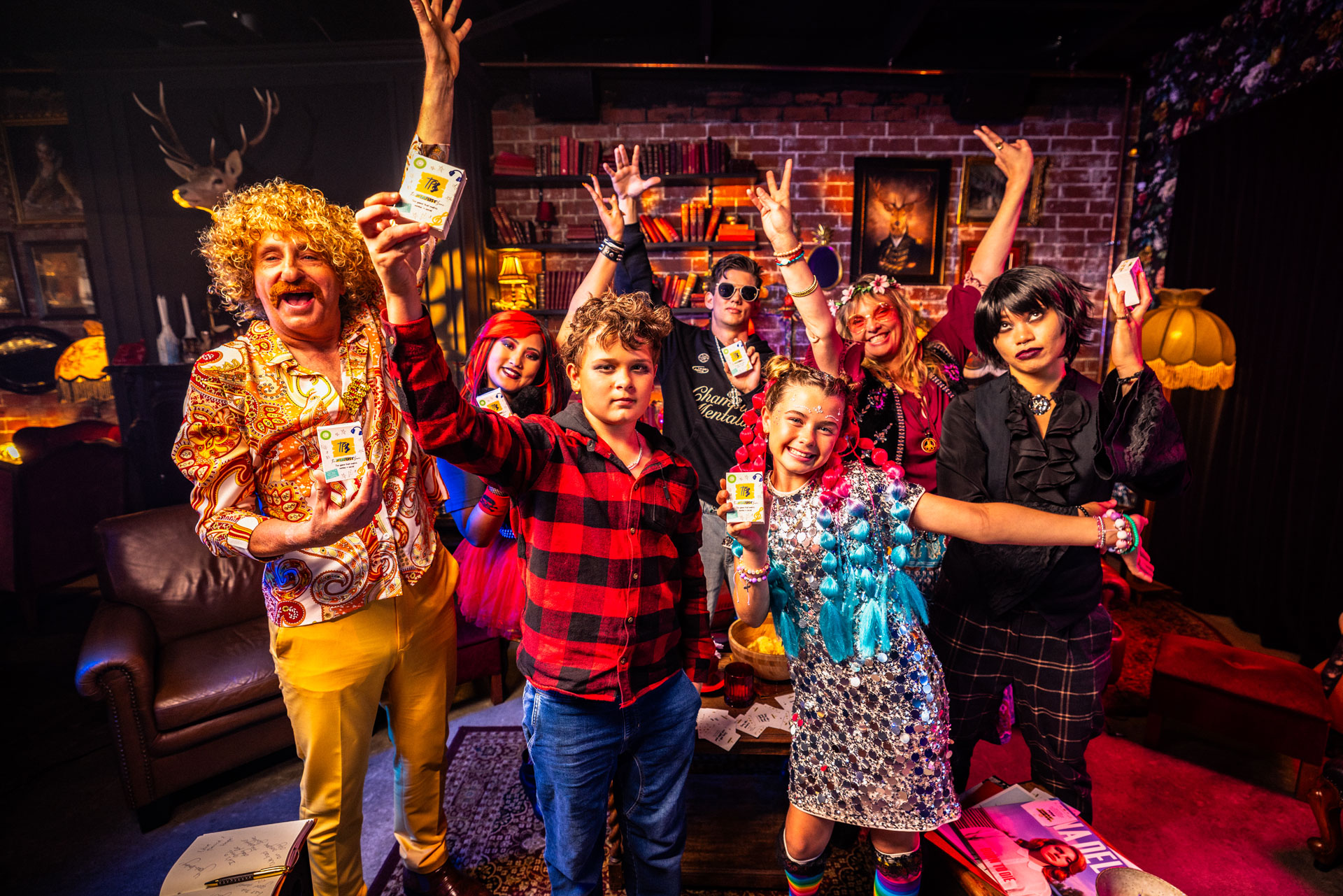 Group of seven people dressed in colourful and eclectic music genre-inspired clothes posing excitedly with their own copies of The Playlist Game.