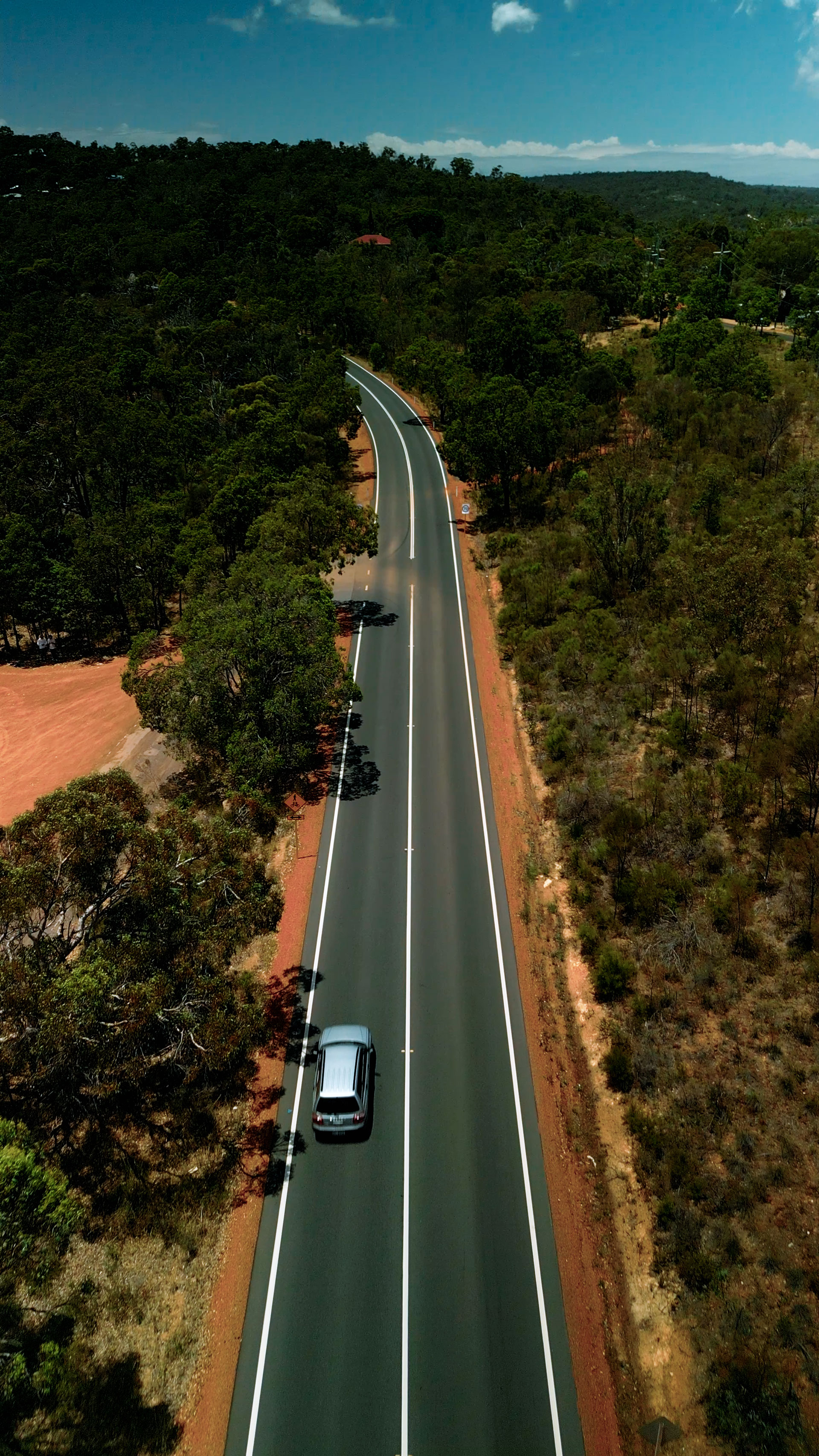 A birds-eye shot of long road in the Australian country surrounded by trees on a sunny day, with a car driving into the distance.