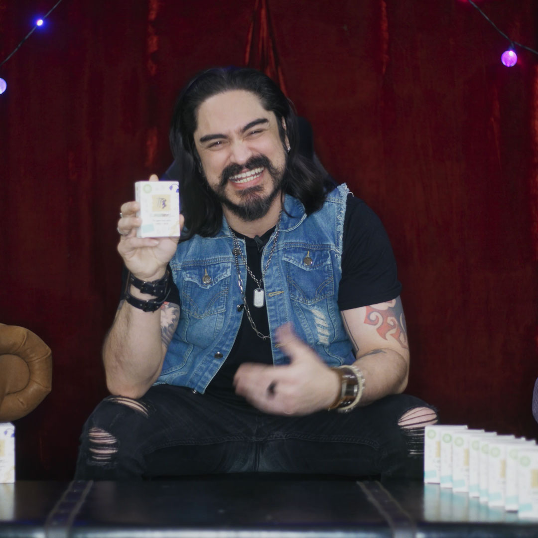 Man with long hair and tattoo on arm smiling and holding a deck of The Playlist Game, sitting in front of a dark red backdrop with string lights and several similar boxed products on the table.