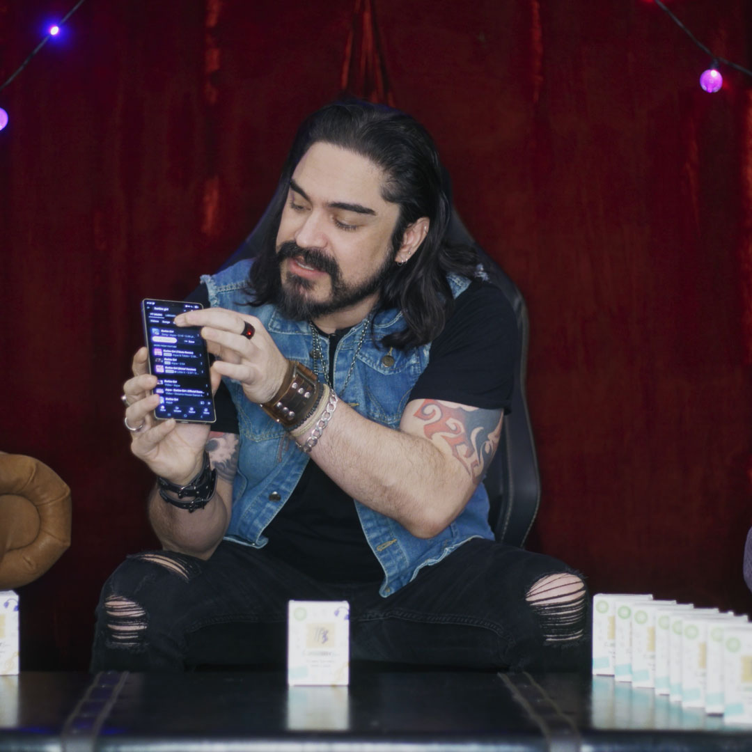 Man with long hair, beard, and tattoos pointing at a smartphone screen while sitting on a chair with product boxes lined up on the table in front.