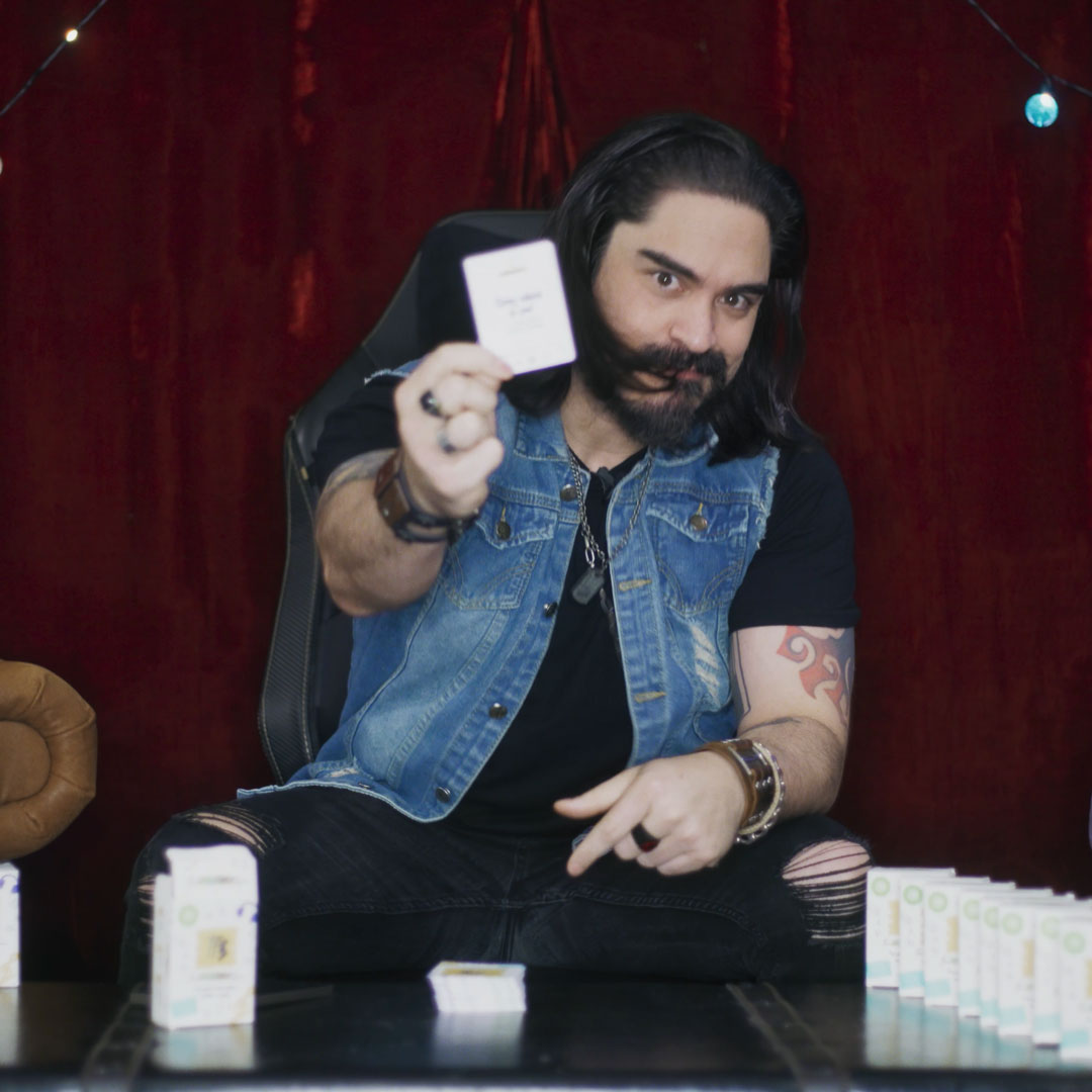 Man with long hair, beard, and tattoos wearing a denim vest holding a card towards the camera while sitting with stacks of cards on the table.