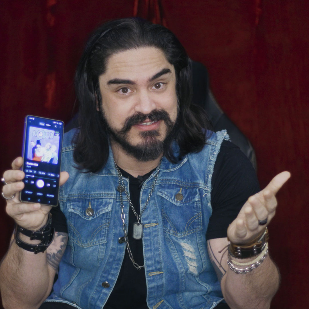Man with long dark hair and beard wearing a denim vest, showing a smartphone with a music app open and gesturing with his other hand.