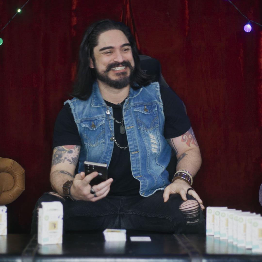 Smiling man with long hair and beard wearing a sleeveless denim vest and black shirt, sitting with tattooed arms holding a smartphone.