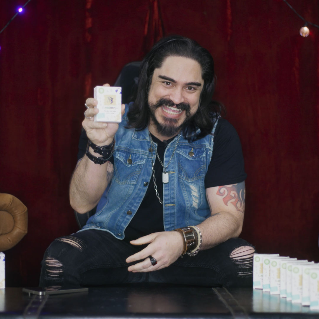 Man with long hair, a beard, and tattoos wearing a denim vest and black shirt, smiling and holding up a small boxed product.
