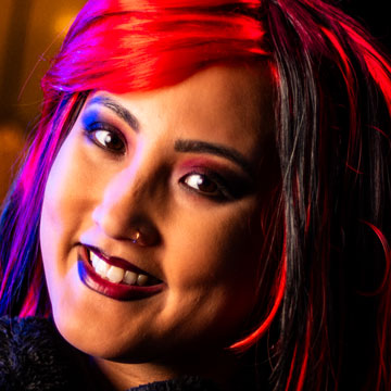 Smiling woman with red and black hair, wearing dark makeup and black earmuffs with a blurred lamp in the background.