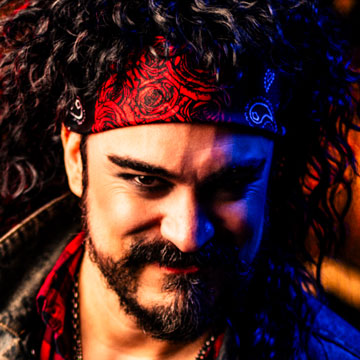 Smiling man with curly hair wearing a red patterned bandana and a beard, lit with blue and warm lighting.