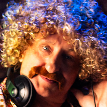 Man with curly blond hair wig, brown mustache, and headphones around his neck.