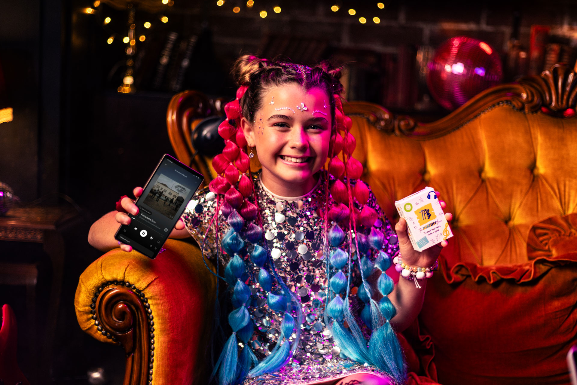 Smiling girl with pink and blue braided hair and sequined dress sitting on an orange vintage sofa, holding a smartphone displaying a music app and a deck of The Playlist Game.