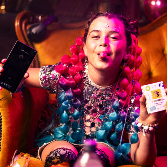 Young woman with pink and blue bubble braids sitting on a yellow chair, sticking out her tongue, holding a smartphone in one hand and a deck of The Playlist Game in the other.