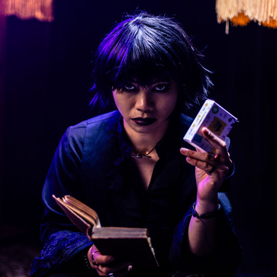 Young woman with dark hair and makeup holding an open book in one hand and a deck of The Playlist Game in the other under purple lighting.