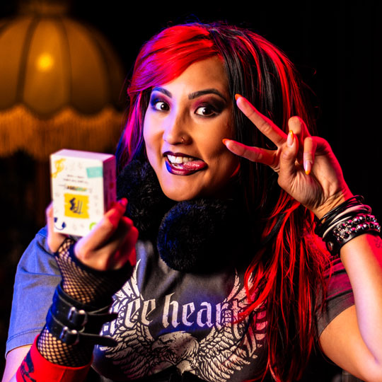 Young woman with red and black hair making a peace sign and holding a deck of The Playlist Game, wearing a graphic tee and fingerless gloves.