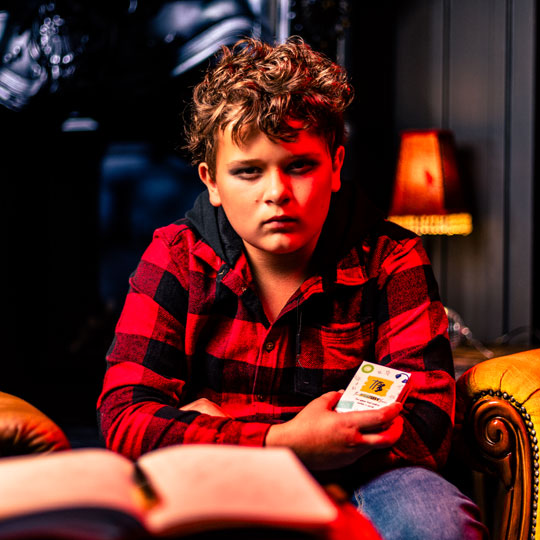 Boy in a red and black plaid shirt sitting on a leather armchair, holding a deck of The Playlist Game with a lamp in the background.