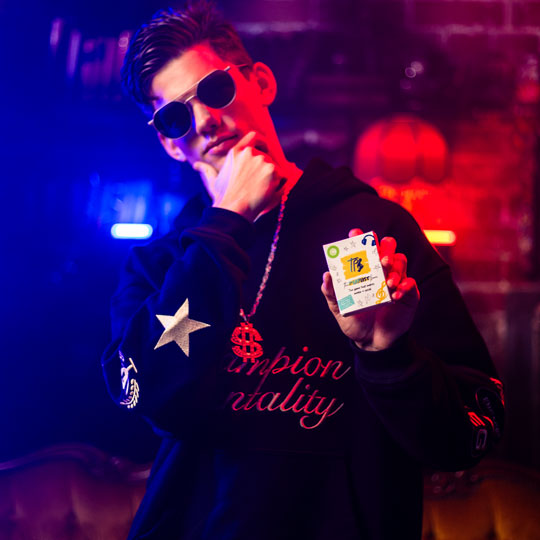 Young man wearing sunglasses and a black hoodie with a dollar sign necklace, holding up the Playlist Game with a confident pose against a colorful neon-lit background.