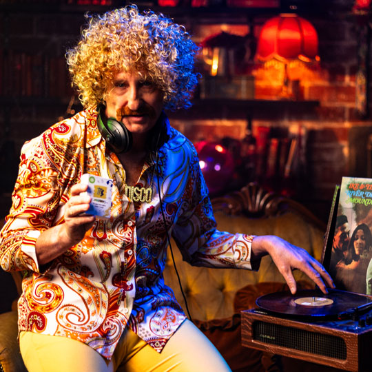 Man with curly blonde hair and patterned shirt holding a deck of The Playlist Game while operating a vintage record player in a cozy, retro room.