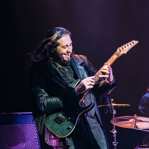 Man with long hair and leather jacket playing electric guitar on stage with drum set in background.