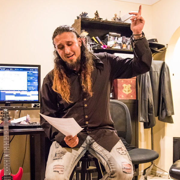 Man with long hair and beard sitting on a stool holding a paper and gesturing with his hand in a room with a guitar, computer, and shelves.