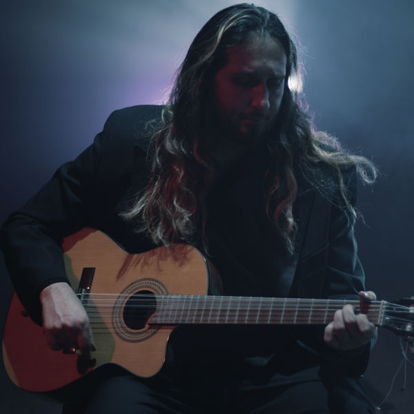 Man with long hair playing an acoustic guitar on stage with moody blue lighting.